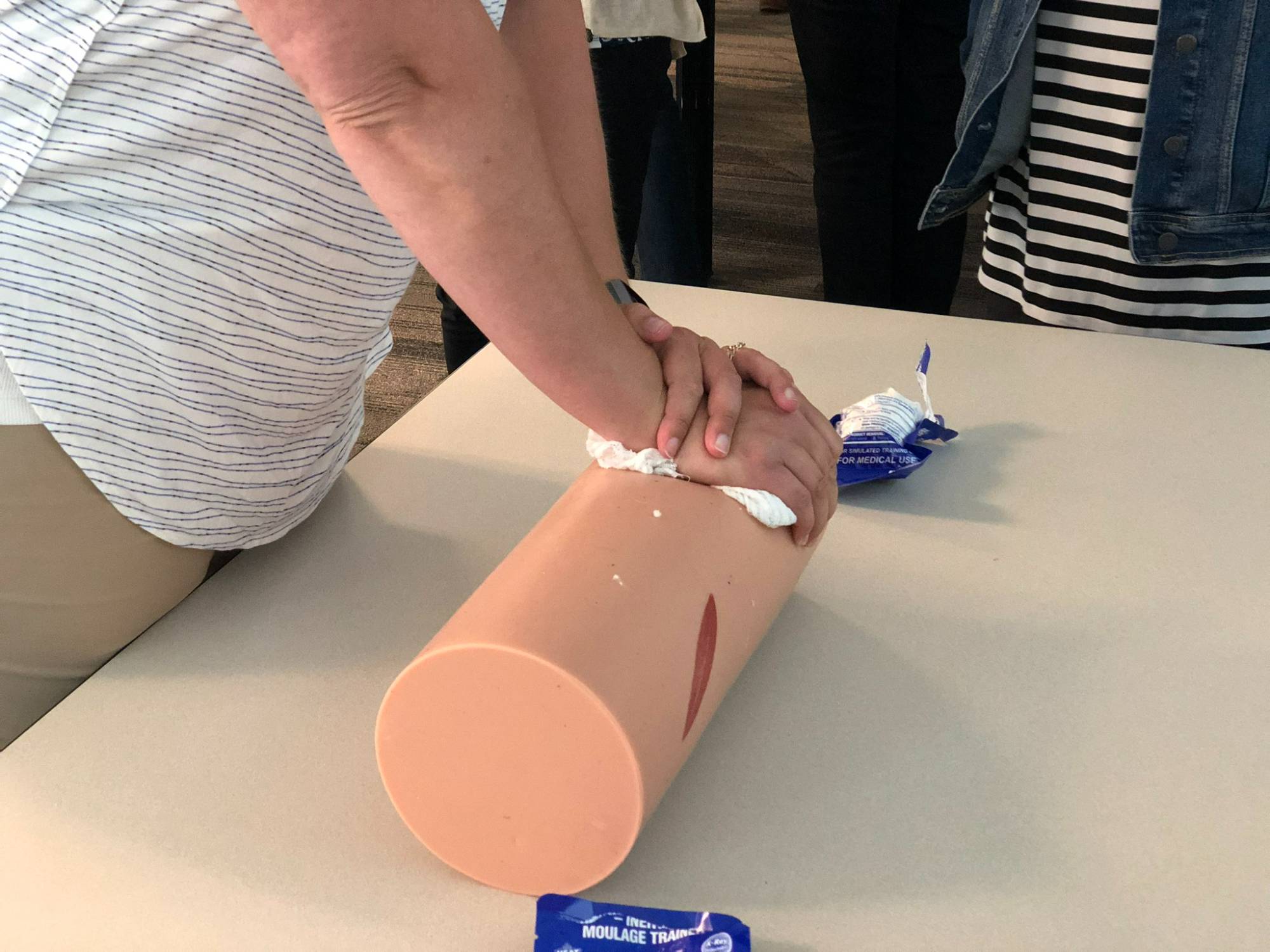 White woman's arms and hands crossed over a cylindrical item meant to simulate a person's leg, packing a wound with gauze